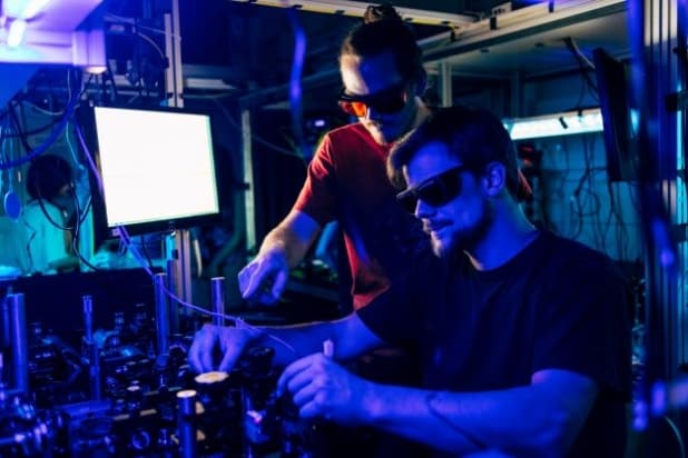 Sebastian Ecker and Martin Bohmann leaning over an optical bench bathed in purple-blue light