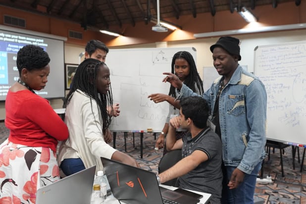 Six people in a classroom sit and stand around a laptop