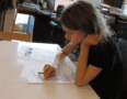 A child sits at a table with a paper in front of her and a desk piled high with papers and books in the background